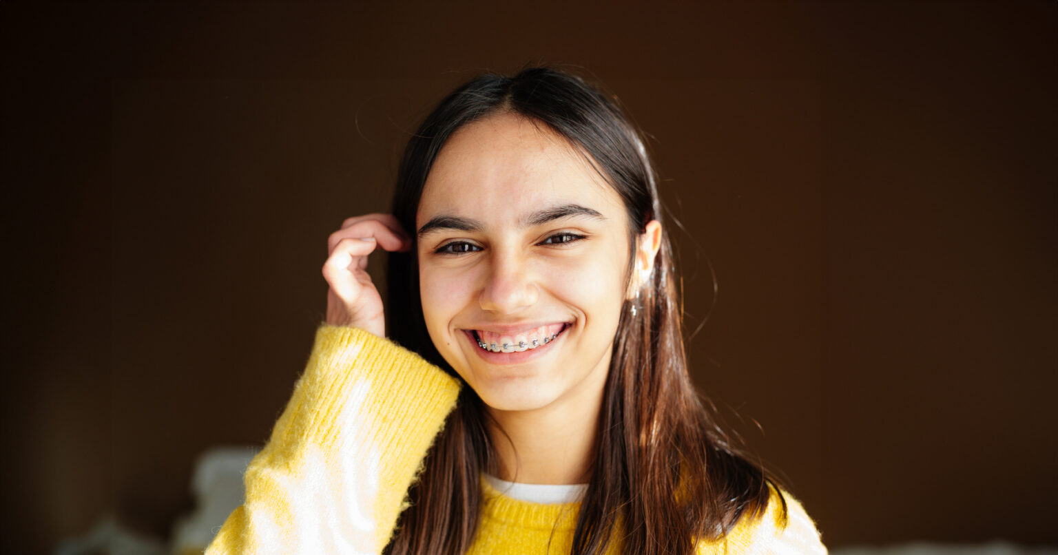 Teen with braces tucking her hair behind her ear and smiling as rays of afternoon sun shine through