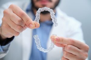 An orthodontist in a white doctor's coat holds up clear aligners to the camera