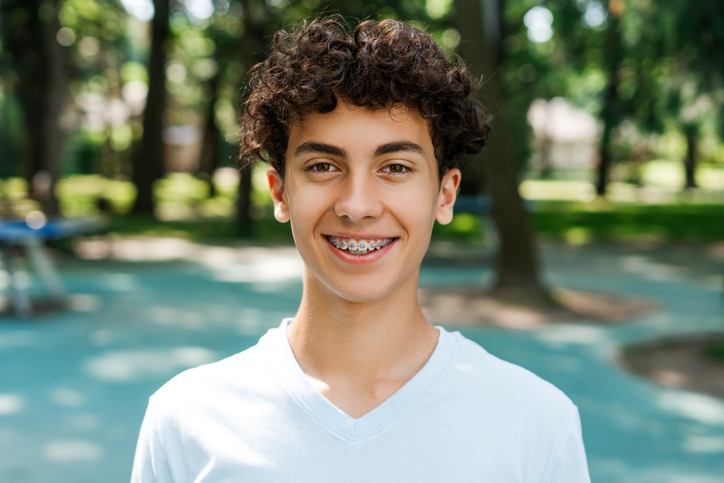 Portrait of positive teenager boy with braces in white t shirt looking at camera at the park.
