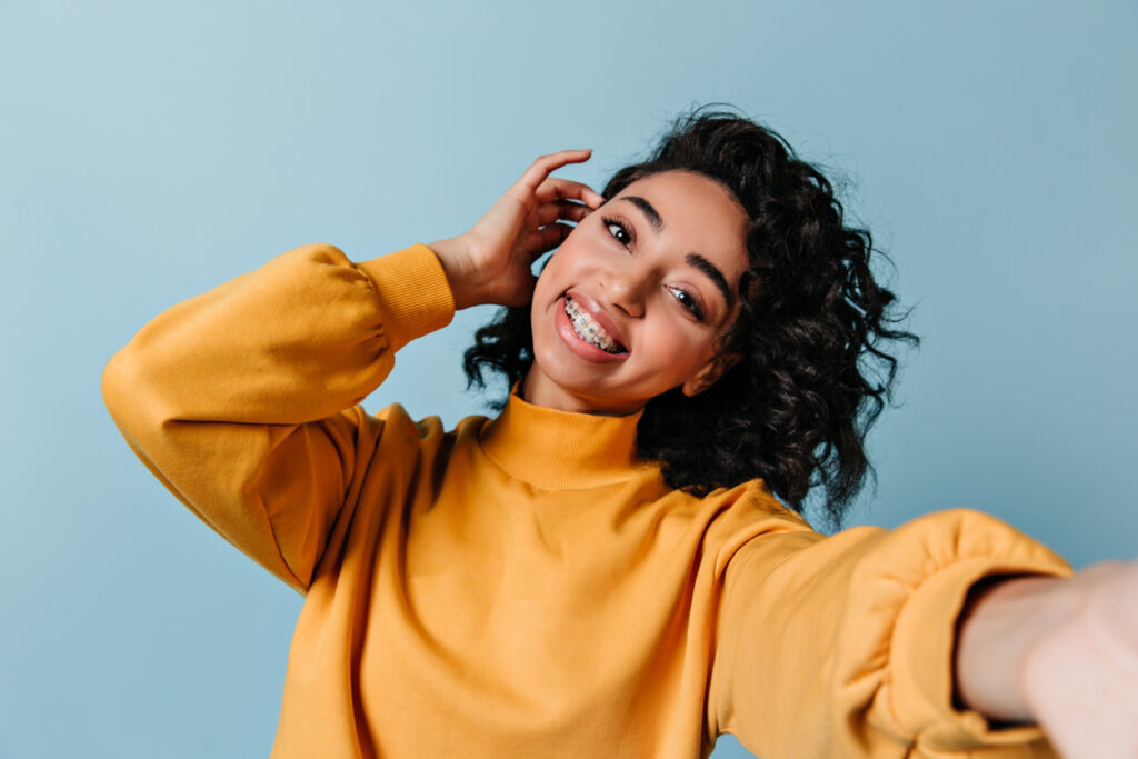Teen with braces and dark curly hair smiling and tucking her hair behind her ear