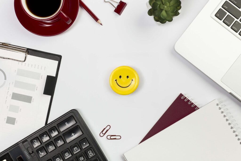 A section of a white desk with a calculator, paper clips, notebooks, and a yellow smiley face