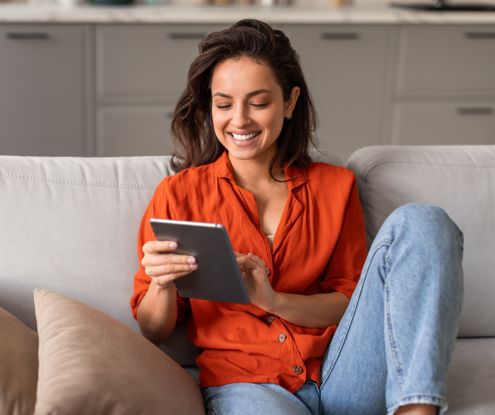 Cheerful young female in vibrant orange blouse engrossed in her tablet while comfortably reclining on chic gray sofa at home, free space